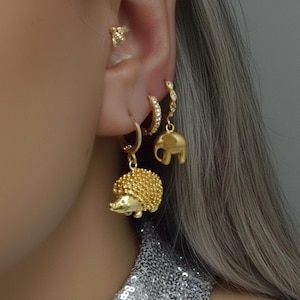 Op de afbeelding: Een close-up van een oor versierd met meerdere gouden oorbellen. De oorbellen omvatten een kleine bijenknop, een hoepel met kleine heldere stenen, een bungelende olifant en een egel. De gouden sieraden voegen een vleugje elegantie toe.