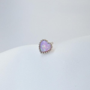 Op de afbeelding: Een enkele oorbel met een hartvormige opaalsteen in roze en lavendel en een zilveren, met kralen bezette rand. De opaalsteen heeft een iriserend uiterlijk. De achtergrond is wit.