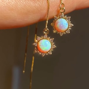 Op de afbeelding: Een paar gouden oorhangers. Elke oorbel heeft een ronde, iriserende opaalsteen, omgeven door een zonnestraalontwerp van kleine, heldere kristallen. De oorbellen hangen aan fijne gouden kettingen.