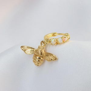 Op de afbeelding: Goudkleurige oorbel met een bijenbedel en een ring versierd met kleine, witte edelstenen. De bij heeft gedetailleerde vleugels en een lichaam, tegen een witte achtergrond.
