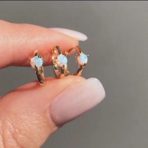 Op de afbeelding: Drie goudkleurige oorringen, elk met een ronde, witte opaalsteen. De oorbellen worden door een hand omhoog gehouden, wat hun ontwerp en grootte laat zien. De oorbellen zijn klein en delicaat.