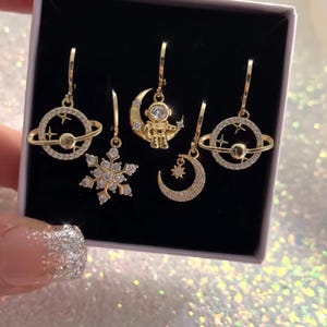 Op de afbeelding: Een set gouden oorhangers met ruimtethema's. Ontwerpen omvatten planeten, een sneeuwvlok, een maan en een astronaut. Elke oorbel is versierd met sprankelende kristallen, gepresenteerd in een zwarte doos.