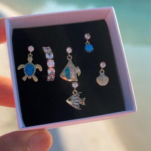 Op de afbeelding: Een verzameling goudkleurige oorbellen en een ring, gepresenteerd in een witte doos met een zwart interieur. De oorbellen hebben verschillende ontwerpen, waaronder een schildpad, een vis en een zeeschelp, met blauwe en heldere edelsteenaccenten. De ring heeft heldere edelstenen.