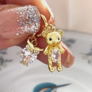 Op de afbeelding: Gouden bedeloorbellen met een teddybeer en een uil. De teddybeer bedel heeft een heldere, ovaalvormige steen. De uil bedel heeft een grote, ronde, heldere steen. De oorbellen worden door een persoon vastgehouden.
