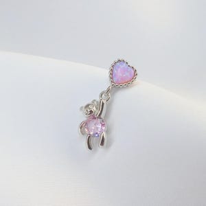 Op de afbeelding: Een zilverkleurige bedel met een kleine teddybeer die een roze hartvormige edelsteen vasthoudt. Het hart heeft een kralenrand en een glanzend, opalescent uiterlijk. De beer heeft een roze edelsteen in zijn buik.