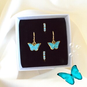 Op de afbeelding: Een sieradenset met goudkleurige vlinder oorbellen en bijpassende oorringen, gepresenteerd in een witte doos met een zwart interieur. De vlindervleugels zijn helder turquoise van kleur, tegen een zachte, witte achtergrond.