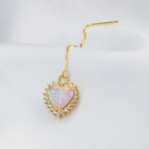 Op de afbeelding: Een gouden oorbel met een hartvormige opalen hanger. De opaal is omgeven door kleine, heldere kristallen en een gouden frame. De oorbel heeft een fijne ketting en een pin.