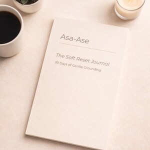 May include: A journal titled "Asa-Ase The Soft Reset Journal 30 Days of Gentle Grounding" lies open on a light surface. A small succulent in a white pot, a cup of coffee, and a lit candle in a glass holder are also present.
