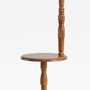 May include: A wooden floor lamp with a round table surface integrated into the central post. The lamp features a beige pleated fabric shade, a turned wood column, and a circular base with small feet. A brown electrical cord is coiled at the base.