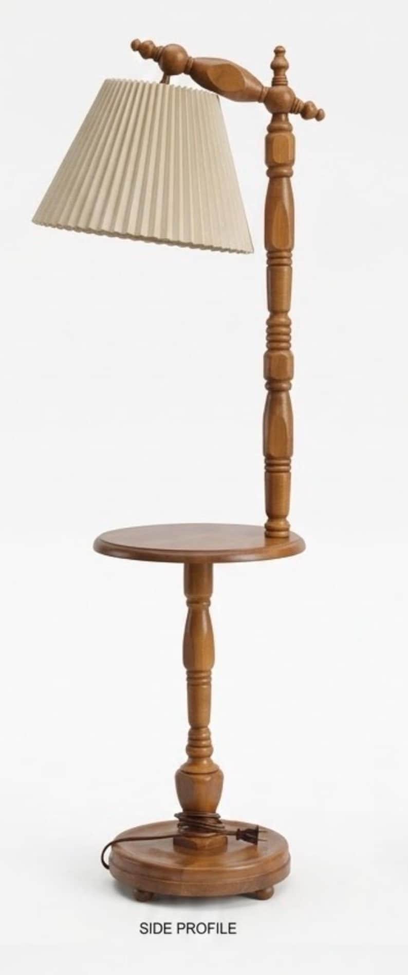 May include: A wooden floor lamp with a round table surface integrated into the central post. The lamp features a beige pleated fabric shade, a turned wood column, and a circular base with small feet. A brown electrical cord is coiled at the base.