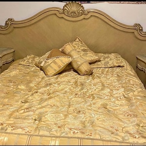 Puede incluir: Un lujoso juego de dormitorio con un cabecero dorado con tallas ornamentadas y mesitas de noche a juego. La cama está cubierta con un edredón y almohadas doradas con estampado floral. Un teléfono antiguo y un pequeño espejo están en las mesitas de noche.