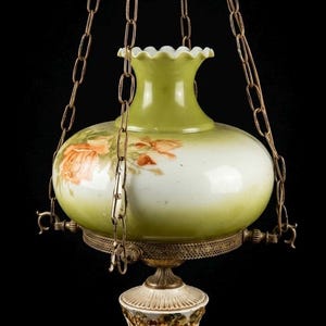 May include: Antique hanging lamp with a green and white glass shade, adorned with orange rose floral designs. The lamp is suspended by brass chains and features a decorative brass base and a detailed bottom fixture.