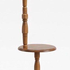 May include: A floor lamp with a wooden stand featuring a round table surface, a central turned wood post, and a beige pleated fabric lampshade. The base is circular with small feet, and a brown electrical cord is coiled at the bottom of the stand.