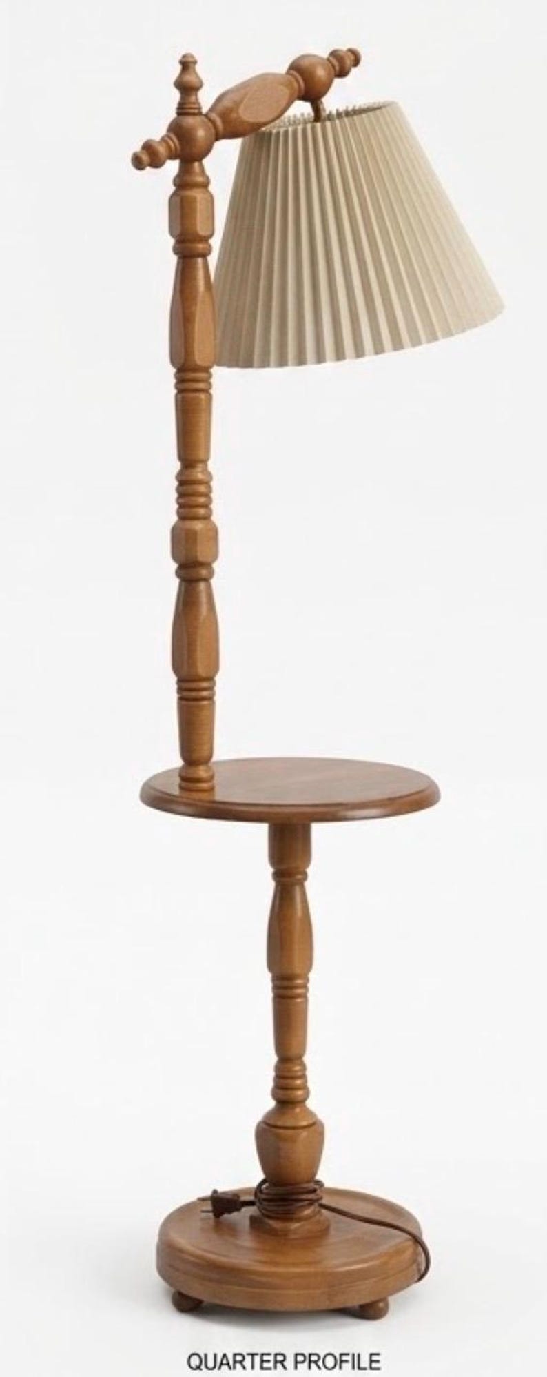 May include: A floor lamp with a wooden stand featuring a round table surface, a central turned wood post, and a beige pleated fabric lampshade. The base is circular with small feet, and a brown electrical cord is coiled at the bottom of the stand.