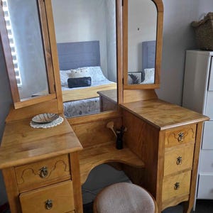 Puede incluir: Tocador de madera con espejo triple, cajones y taburete. El tocador de madera clara tiene tiradores de cajón ornamentados. El taburete tiene un asiento redondo y acolchado. Un plato decorativo está sobre el tocador.