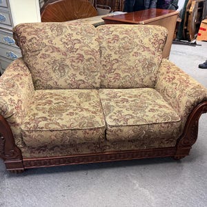 Op de afbeelding: Tweezitsbank met een bloemmotief in beige, bruin en rood. De bank heeft een houten frame met sierlijke details op de armleuningen en de basis. De kussens zijn zacht en uitnodigend, perfect voor een woonkamer.