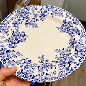 Puede incluir: Un plato de cerámica blanco con un diseño floral azul. El plato presenta un patrón detallado de flores y hojas azules alrededor del borde y en grupos en la superficie. El plato tiene un borde festoneado.
