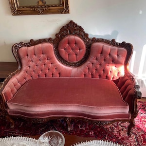 May include: A dark wood settee featuring a curved backrest with intricate carved details and a central circular tufted panel. The seat and back are upholstered in a dusty rose velvet fabric with button tufting throughout the backrest area.
