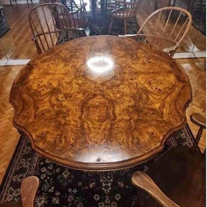May include: A round wooden dining table featuring a prominent burl wood grain pattern with a scalloped edge. The table surface displays a rich, swirling amber and brown finish, surrounded by several wooden spindle-back chairs in a room setting.