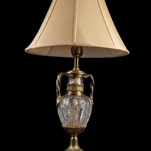 May include: A decorative table lamp with a beige lampshade and a clear, patterned glass base. The base has brass accents and two twisted handles. The lamp sits on a brass base and has a black background.