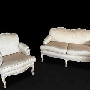 May include: A cream-colored armchair and a matching two-seater sofa featuring a subtle, swirling tonal pattern. Both pieces have curved backrests, rolled arms, and light-colored wooden cabriole legs with a distressed finish on the frame.