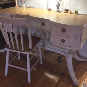 May include: A white desk with two drawers and a matching chair. The desk has a light wood top and curved edges. The chair has a wooden seat and white painted frame. The desk and chair are set on a wooden floor.