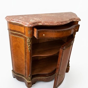 May include: A wooden cabinet featuring a pink marble top, one pull-out drawer, and a single hinged door. The exterior displays intricate wood grain patterns, gold-toned metal decorative accents, and ornate metal hardware on the drawer and door.