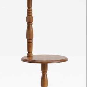 May include: A floor lamp with a round wooden side table integrated into its central post. The lamp features a turned wood stand, a circular base, and a beige pleated fabric lampshade. A brown electrical cord is coiled at the base of the lamp.