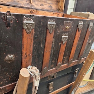 May include: An antique trunk with a black exterior, accented by wooden slats and decorative metal hardware. The trunk has a lock and is open, revealing the interior. The trunk is likely used for storage and travel.