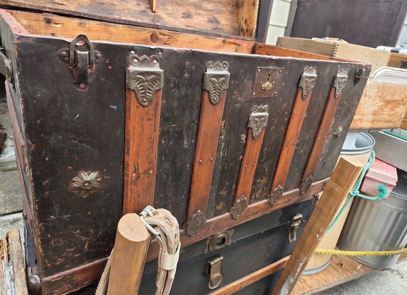 May include: An antique trunk with a black exterior, accented by wooden slats and decorative metal hardware. The trunk has a lock and is open, revealing the interior. The trunk is likely used for storage and travel.