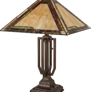 May include: A bronze-colored table lamp with a square stained-glass shade. The shade features a geometric design with beige, purple, and green glass panels. The lamp has a rectangular base and a vertical support structure.