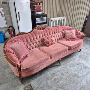 Op de afbeelding: Een vintage roze fluwelen bank met een gecapitonneerde rugleuning en drie zitkussens. De bank heeft een gebogen houten frame en is versierd met drie decoratieve kussens. De kussens zijn roze en hebben franjes.
