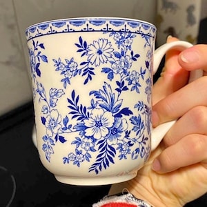 May include: A cream-coloured ceramic mug adorned with a blue floral pattern. The mug has a handle and a detailed design of flowers and leaves. The rim of the mug has a decorative blue border. The mug is held by a person.
