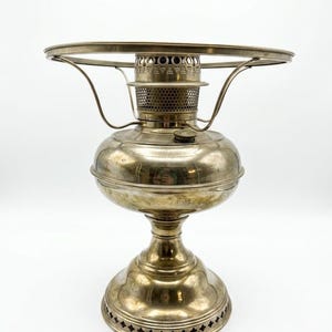 May include: A vintage silver-toned oil lamp. It has a round base and a bulbous body. The lamp features a decorative top with a circular handle and a mesh section around the burner. The base has a detailed, patterned design.
