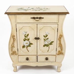 Op de afbeelding: Crèmekleurige kast met bloemendetails. De kast heeft twee deuren, twee lades en een bovenste lade. De deuren en de bovenkant zijn versierd met geschilderde witte bloemen en groene bladeren. Goudkleurige decoratieve accenten bevinden zich aan de zijkanten.