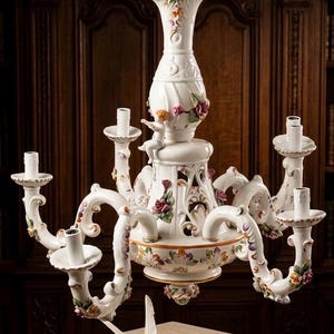 May include: A white porcelain chandelier featuring five candle holders, ornate floral motifs, and a small cherub figure. The central column is topped with a quill and inkwell design. A matching small inkwell with a quill and a miniature book sit below.