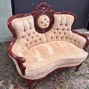 May include: A loveseat featuring a dark mahogany-coloured wooden frame with intricate carved details and a central circular tufted accent. The seat and backrest are upholstered in a light beige, textured fabric with deep button tufting.