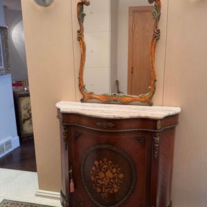 May include: An antique wooden cabinet with a marble top, ornate carvings, and a floral inlay. A decorative mirror with a gold and green frame hangs above the cabinet. The cabinet has a key and a pink tassel.