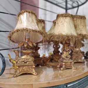 Puede incluir: Un conjunto de lámparas ornamentadas de estilo antiguo con bases doradas y pantallas color crema. Las pantallas están adornadas con un material con flecos de color beige. Las lámparas están dispuestas sobre una mesa redonda de color claro.