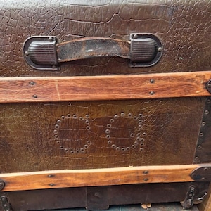 Puede incluir: Ba&uacute;l marr&oacute;n vintage con exterior texturizado, detalles de madera y herrajes met&aacute;licos. El ba&uacute;l presenta un asa de cuero y las iniciales "CG" delineadas con remaches. Una pieza de equipaje cl&aacute;sica.