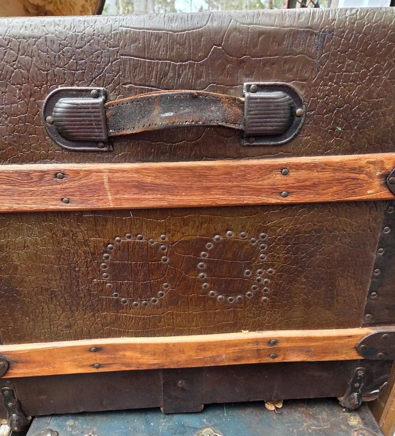 Puede incluir: Ba&uacute;l marr&oacute;n vintage con exterior texturizado, detalles de madera y herrajes met&aacute;licos. El ba&uacute;l presenta un asa de cuero y las iniciales "CG" delineadas con remaches. Una pieza de equipaje cl&aacute;sica.