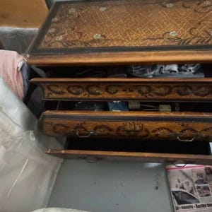 Può includere: Un comò in legno con tre cassetti, con intagli floreali ornati e una finitura marrone scuro. La superficie superiore presenta un motivo intrecciato. I cassetti sono parzialmente aperti, rivelando l'interno.