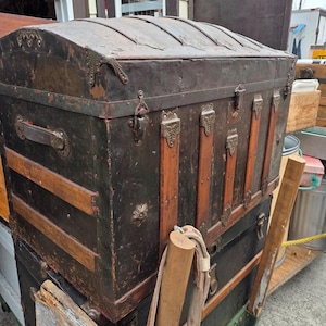 May include: An antique black trunk with a curved top and metal hardware. The trunk has wooden slats and decorative metal accents. The trunk is stacked on top of another trunk. The trunk is weathered and shows signs of age.