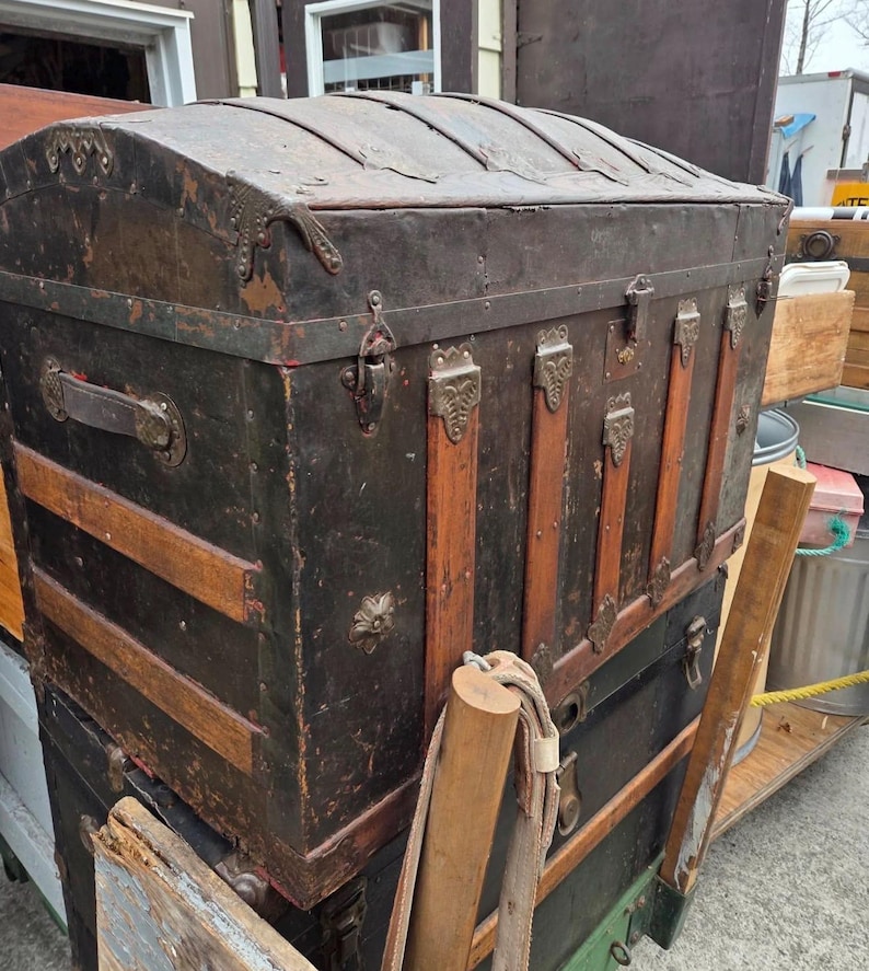 May include: An antique black trunk with a curved top and metal hardware. The trunk has wooden slats and decorative metal accents. The trunk is stacked on top of another trunk. The trunk is weathered and shows signs of age.
