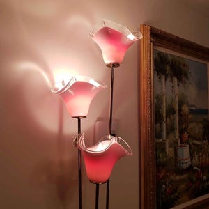 May include: A three-light floor lamp with pink, flower-shaped glass shades. The shades have a ruffled edge and are mounted on slender, dark metal stems. The lamp is positioned next to a framed painting with a gold frame.
