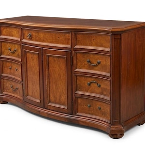Puede incluir: Una cómoda grande de madera con frente curvo y acabado marrón oscuro. Cuenta con múltiples cajones y puertas de armario, con herrajes ornamentados y molduras decorativas. Es un mueble clásico.