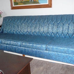 May include: A long, three-cushion sofa upholstered in a vibrant blue fabric with a subtle, repeating floral pattern. The backrest features a distinct diamond-tufted design, and the piece is supported by tapered wooden legs and dark wood armrests.
