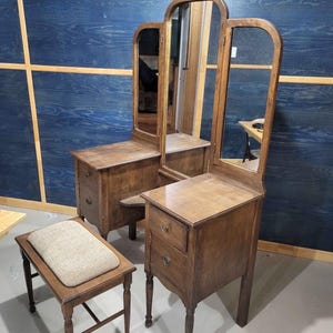 May include: A vintage wooden vanity set with a tri-fold mirror, two small drawers, and a matching stool with a cushioned seat. The vanity and stool are made of dark wood and have a classic design.