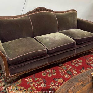 May include: An antique velvet sofa with a dark brown wooden frame. The sofa has three seat cushions and a curved back. The cushions are olive green with pink piping, and the frame has ornate carvings. The sofa sits on a red floral rug.