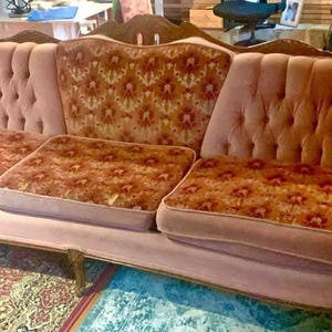 May include: A vintage, three-cushion sofa with a curved back and armrests. The sofa is upholstered in a dusty rose fabric with button tufting on the back and arms. The cushions feature a floral pattern in shades of brown and orange.